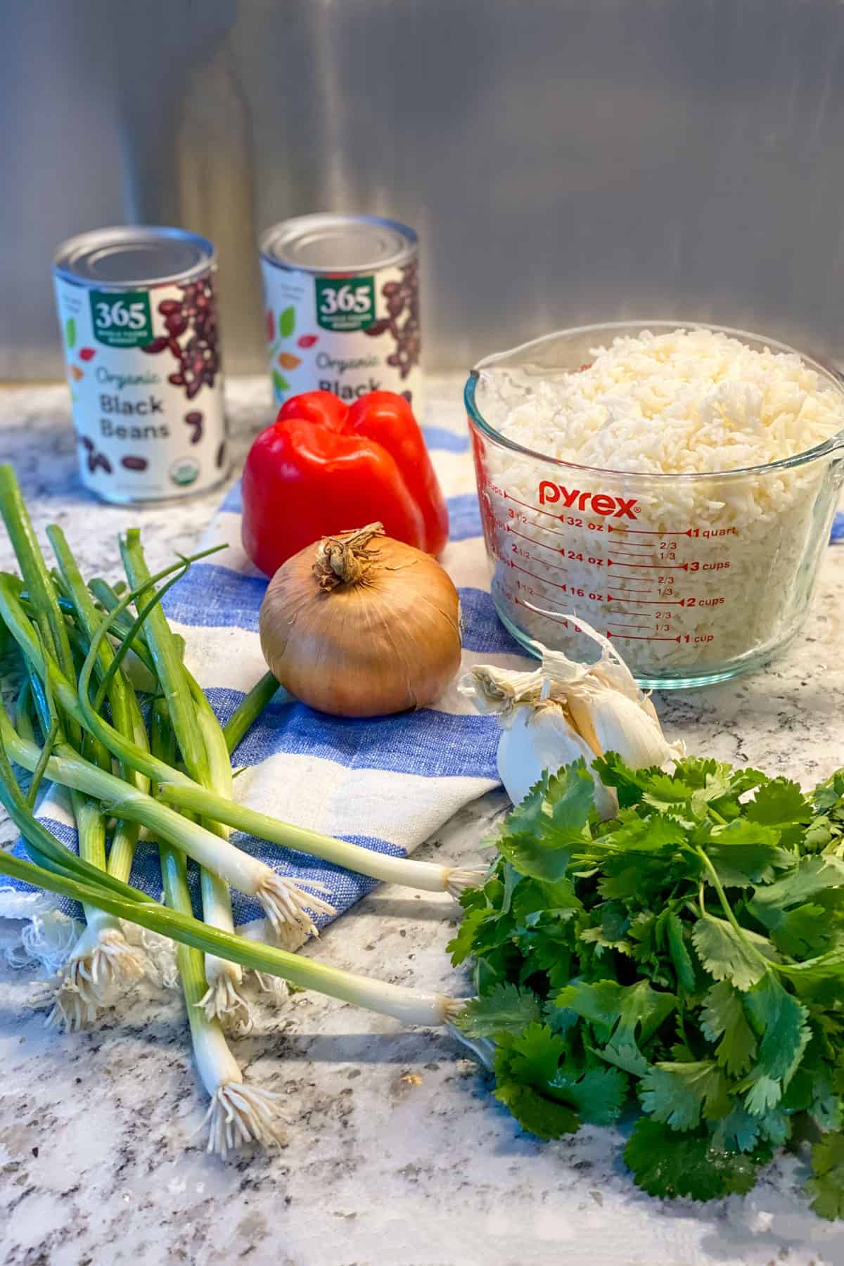 ingredients for gallo pinto, on a white marble counter: a red pepper, bunch of cilantro, bunch of scallions, yellow onion, 2 cans of black beans, measuring cup filled with 6 cups of cooked white rice, head of garlic