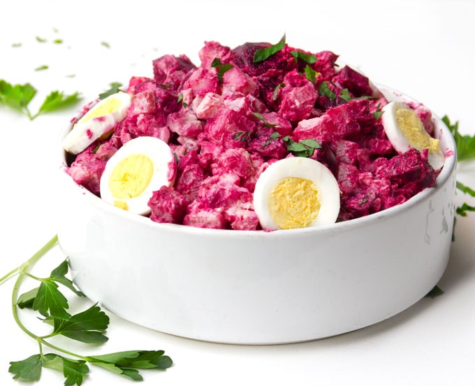 Hot pink Rosolje: Estonian Potato and Beet Salad, in a white bowl, garnished with sliced hard boiled eggs and sprigs of parsley