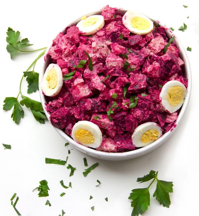 a bowl of hot pink potato salad with slices of hard boiled egg around the rim and garnishes of parsley on the white table surrounding