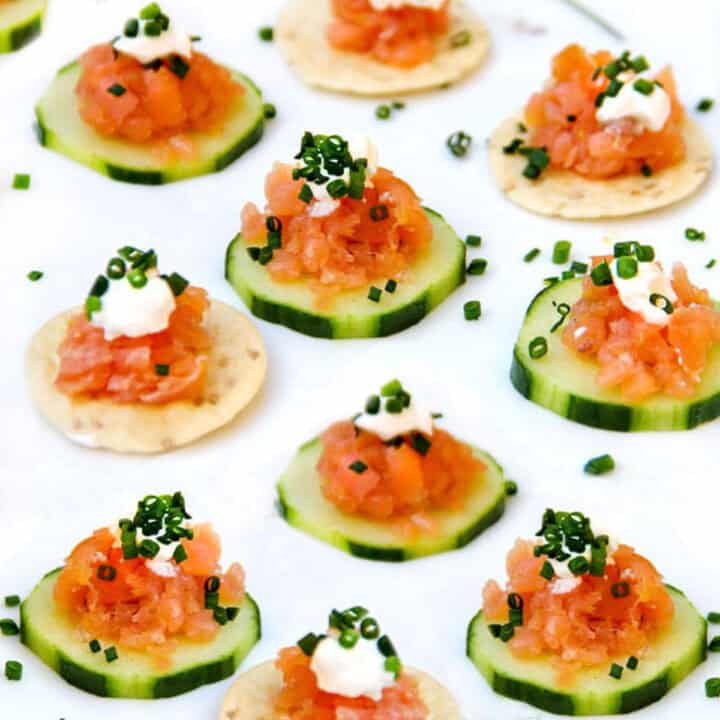 9 little smoked salmon appetizers on a platter with chopped smoked salmon atop cucumber rounds and rice crackers.