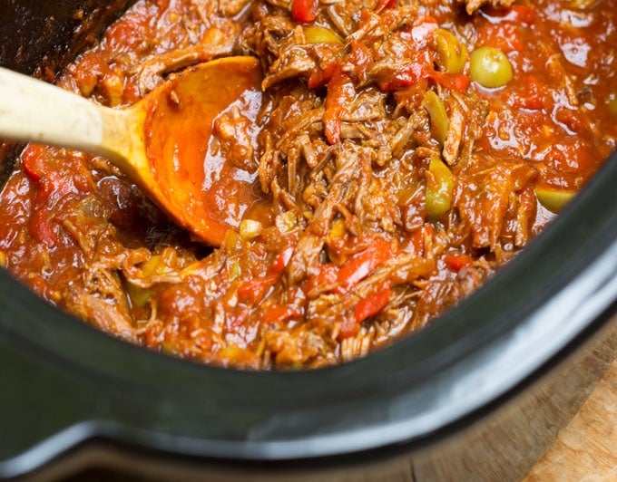 Overhead shot of a slow cooker filled with Cuban ropa vieja (shredded beef stew), a wooden spoon scooping some.