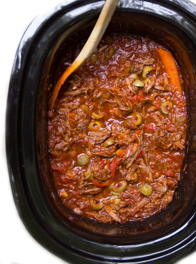 Overhead shot looking into a black oval slow cooker that's filled with Ropa Vieja: Cuban shredded beef stew in tomato sauce with red bell peppers and sliced green olives