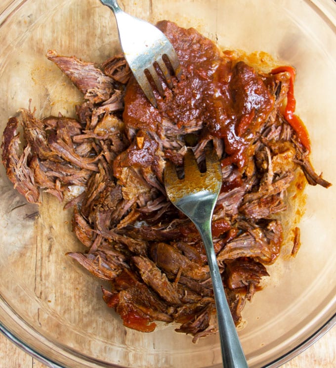 Close up overhead shot of saucy hunks of cuban ropa vieja in a glad bowl, being shredded using two forks