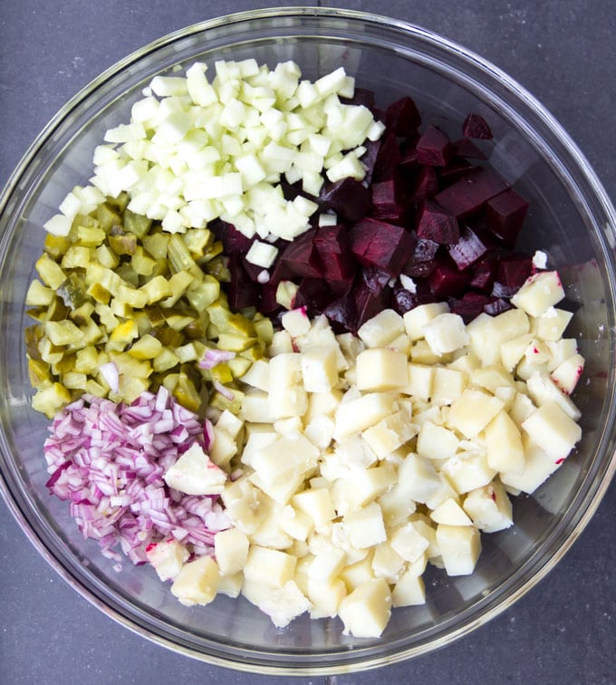 Glass mixing bowl filled with ingredients for Rosolje: a pile of chopped apples, pickles, onions, diced beets and diced potatoes