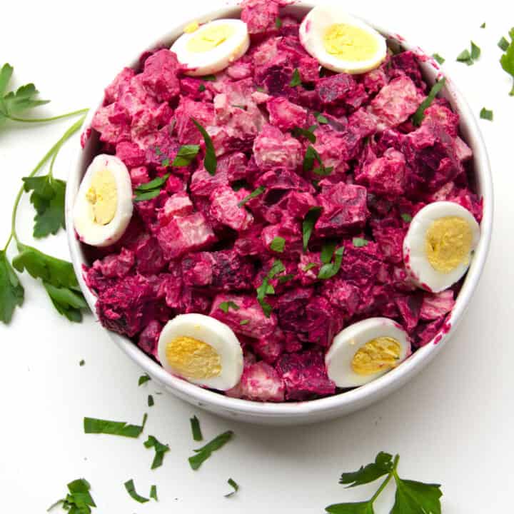 Round serving bowl filled with hot pink rosolje potato salad, with slices of hard boiled egg around the edge.