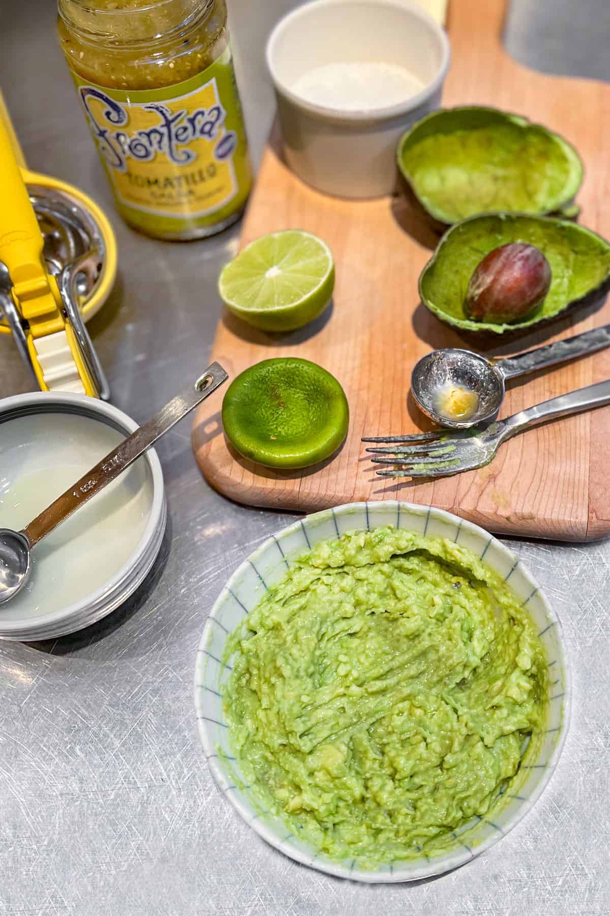 Overhead shot of a small bowl of avocado lime salsa surrounded the empty avocado skin and the pit, the squeezed lime half, and an open jar of salsa verde