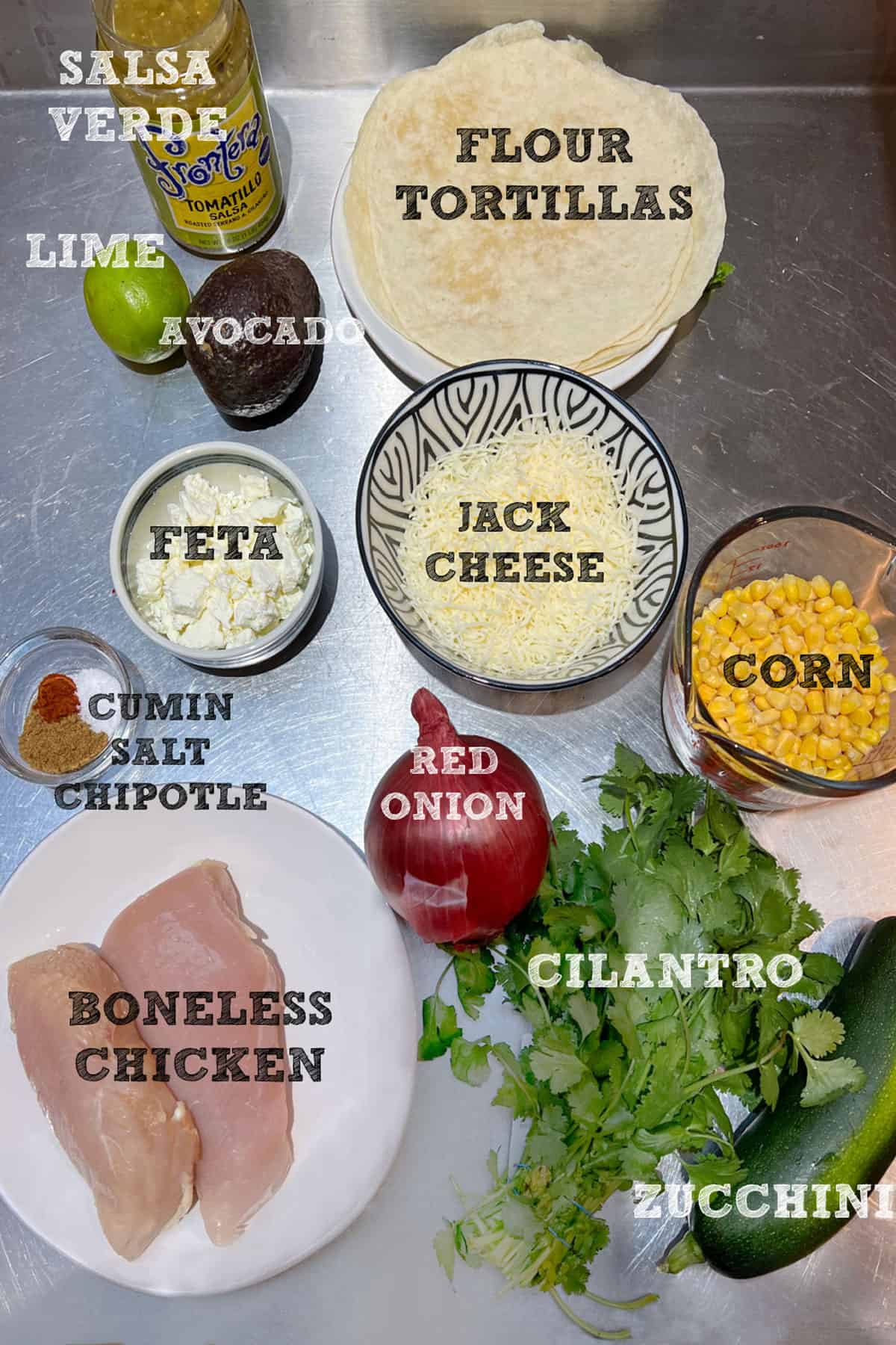 overhead shot of tostada ingredients with labels: tortillas, 2 boneless chicken breasts, a bunch of cilantro, a bowl of shredded jack cheese, a bowl of crumbled feta, a cup of corn kernals, an avocado, a red onion, a lime and a small class bowl with spices measured out