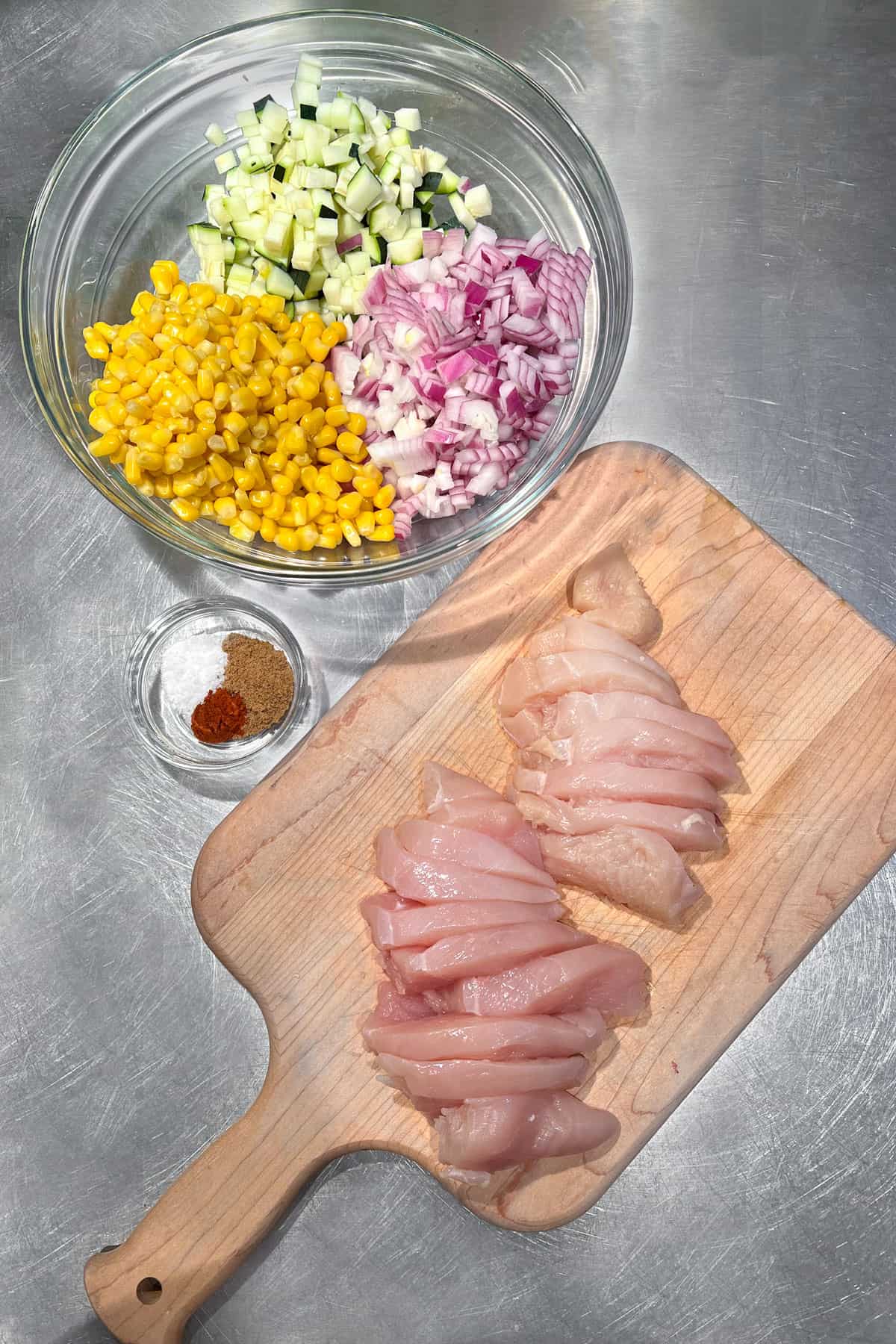 overhead shot of 2 thinly sliced chicken breasts on a wood cutting board. Also a large class bowl filled with a pile of corn, a pile of chopped red onions and a pile of chopped zucchini, also a small bowl of spices