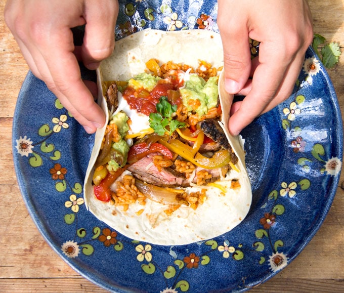 hands folding in the sides of a flour tortilla containing beef, peppers, salsa, sour cream, yellow rice and guacamole