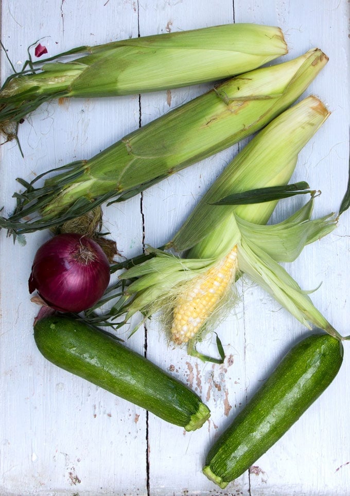 3 cobs of corn, two zucchinis and a red onion on a light blue distressed wooden table