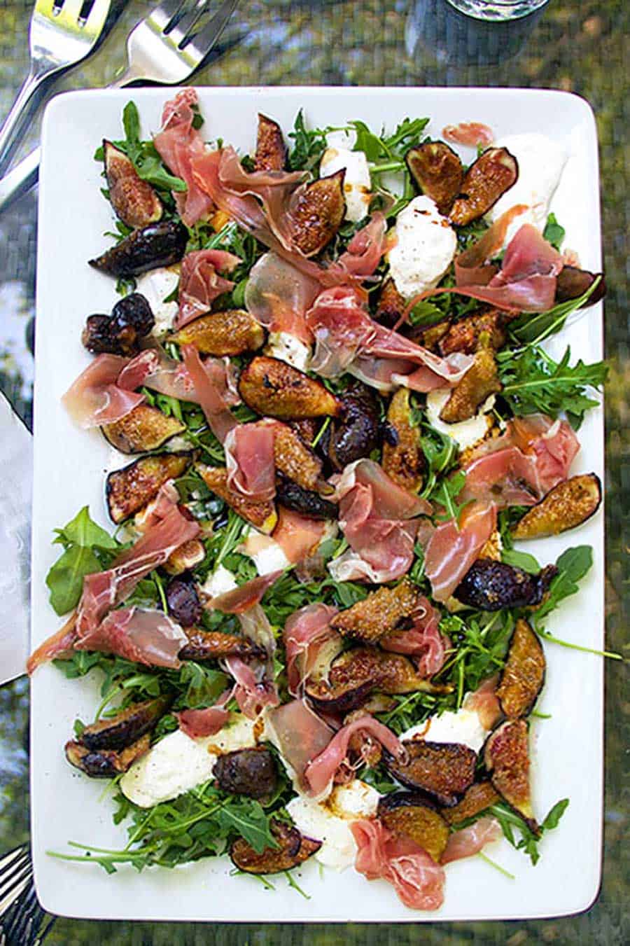 grilled figs, prosciutto and burrata on a large white rectangular platter with some arugula leaves
