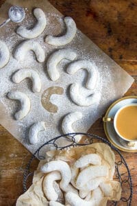 overhead view showing about a dozen almond crescent cookies on a piece of white parchment plus a basket of crescent cookies and a cup of coffee to the side.
