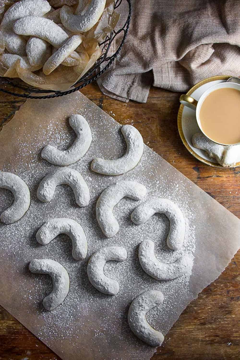 about a dozen sugar-dusted almond crescent cookies on a piece of white parchment paper with a cup of coffee to the side and a basket of more almond crescent cookies 