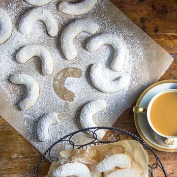 overhead view showing about a dozen almond crescent cookies on a piece of white parchment plus a basket of crescent cookies and a cup of coffee to the side.