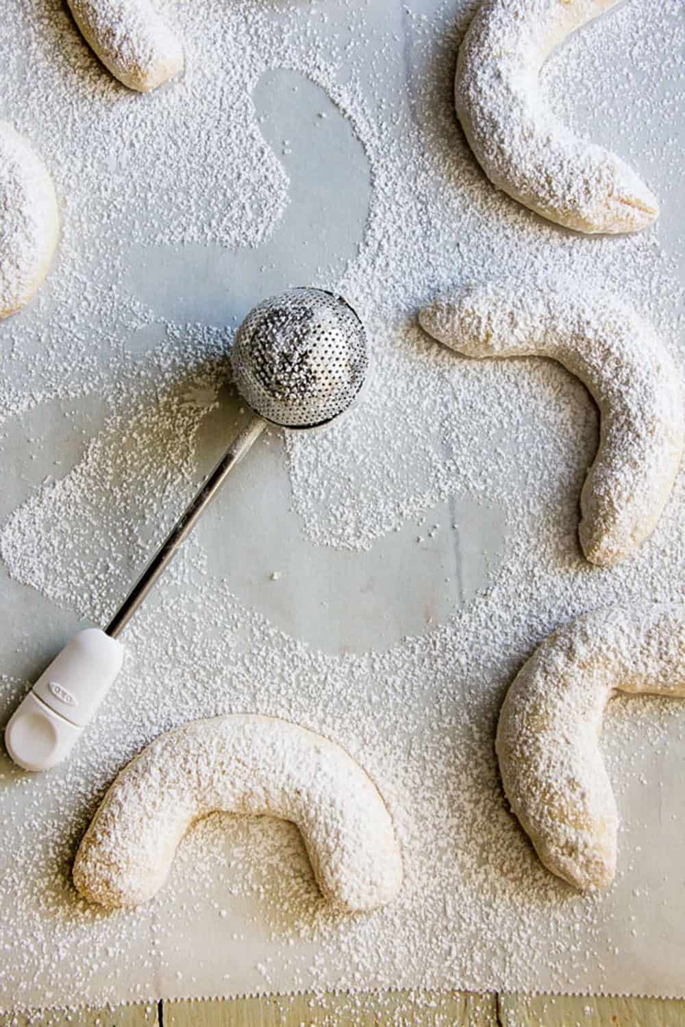 6 almond crescent cookies on a piece of parchment paper with a metal sugar duster
