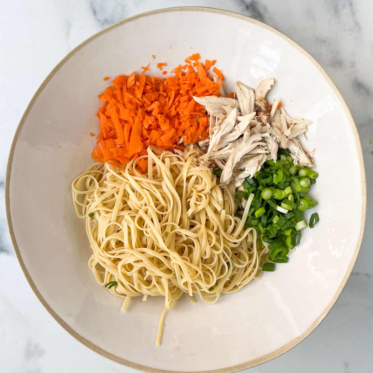 Large white porcelain mixing bowl filled will a pile of cooked linguini, pile of chopped carrots, pile of sliced scallions and pile of shredded cooked chicken.