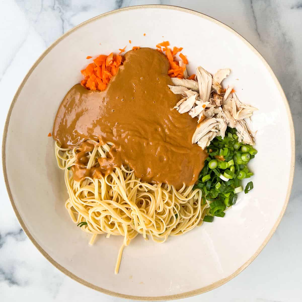 Large white porcelain mixing bowl filled with cooked linguine, chopped carrots, shredded chicken and sliced scallions, all mostly covered with creamy peanut sauce