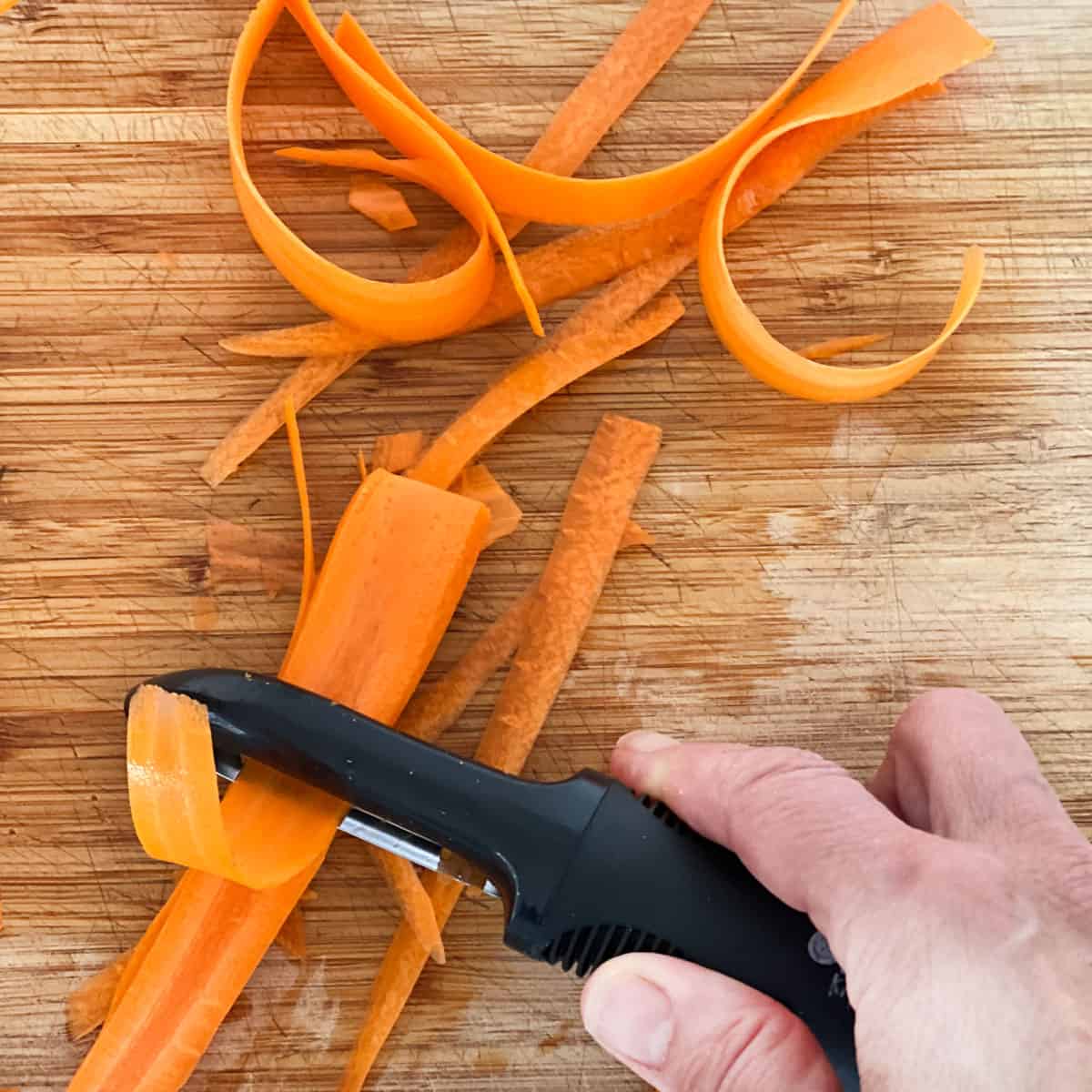 A carrot on a wooden cutting board getting shaved into wide strips with a vegetable peeler.