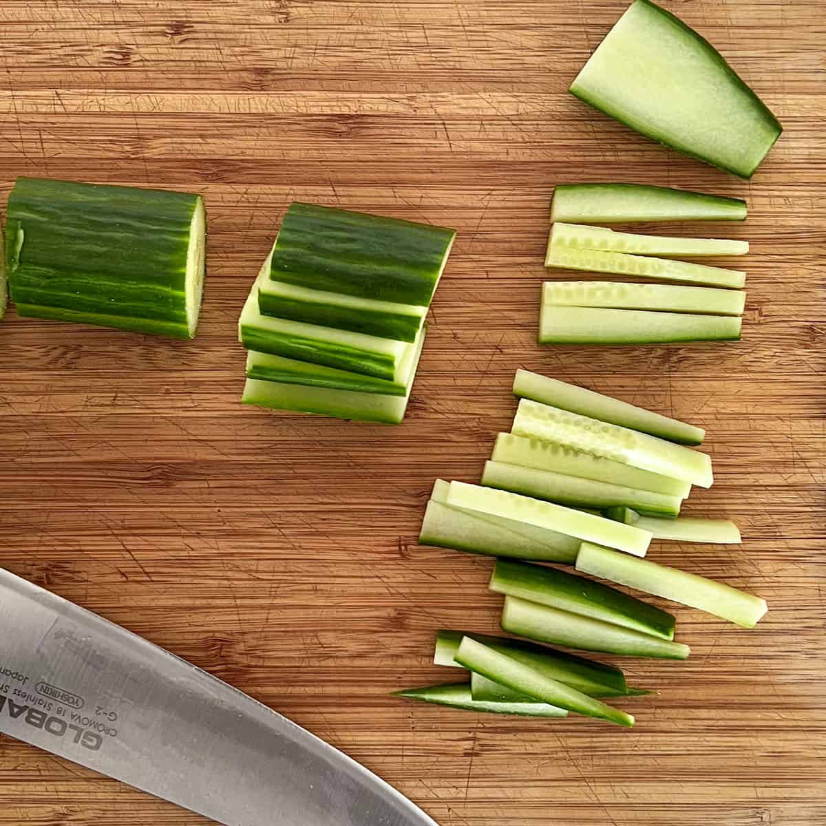 A wooden cutting board topped with segments of English cucumber at different stages of being cut into matchsticks.