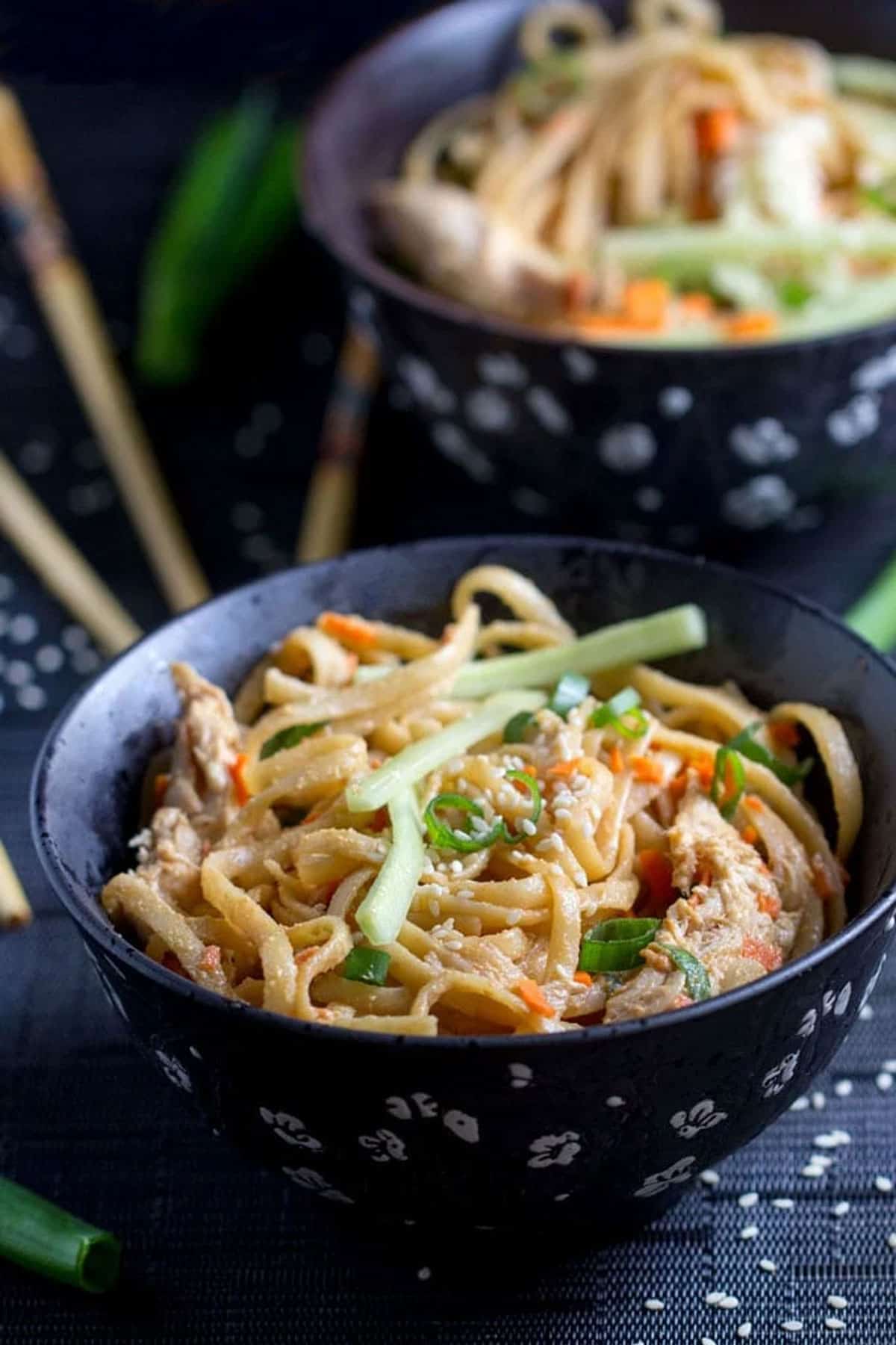 two black bowls filled with linguine coated with peanut sauce, bits of chopped carrots, scallions and sliced cucumbers.