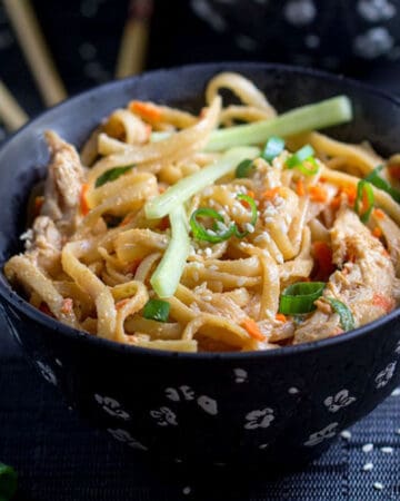 bowl filled with sesame coated linguine noodles, topped with matchstick cucumbers and carrots, sliced scallions and sesame seeds.