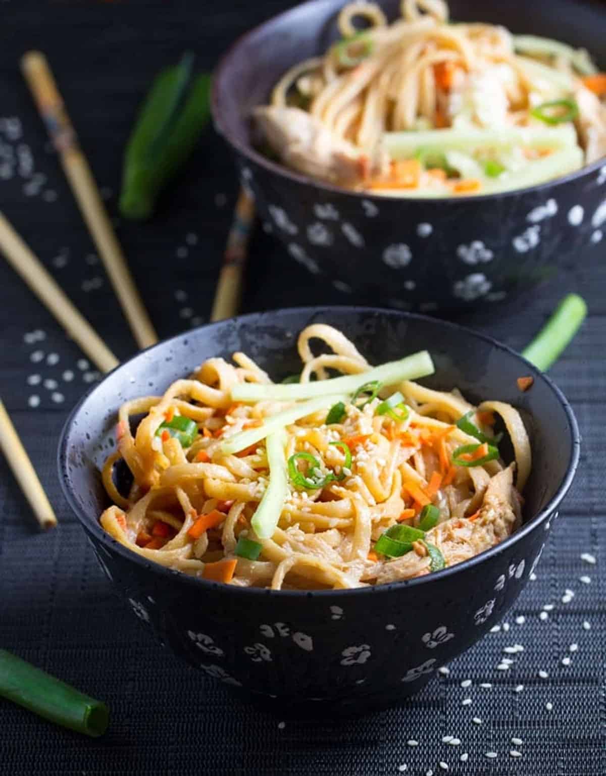 two black bowls filled with linguine coated with peanut sauce, bits of chopped carrots, scallions and sliced cucumbers.