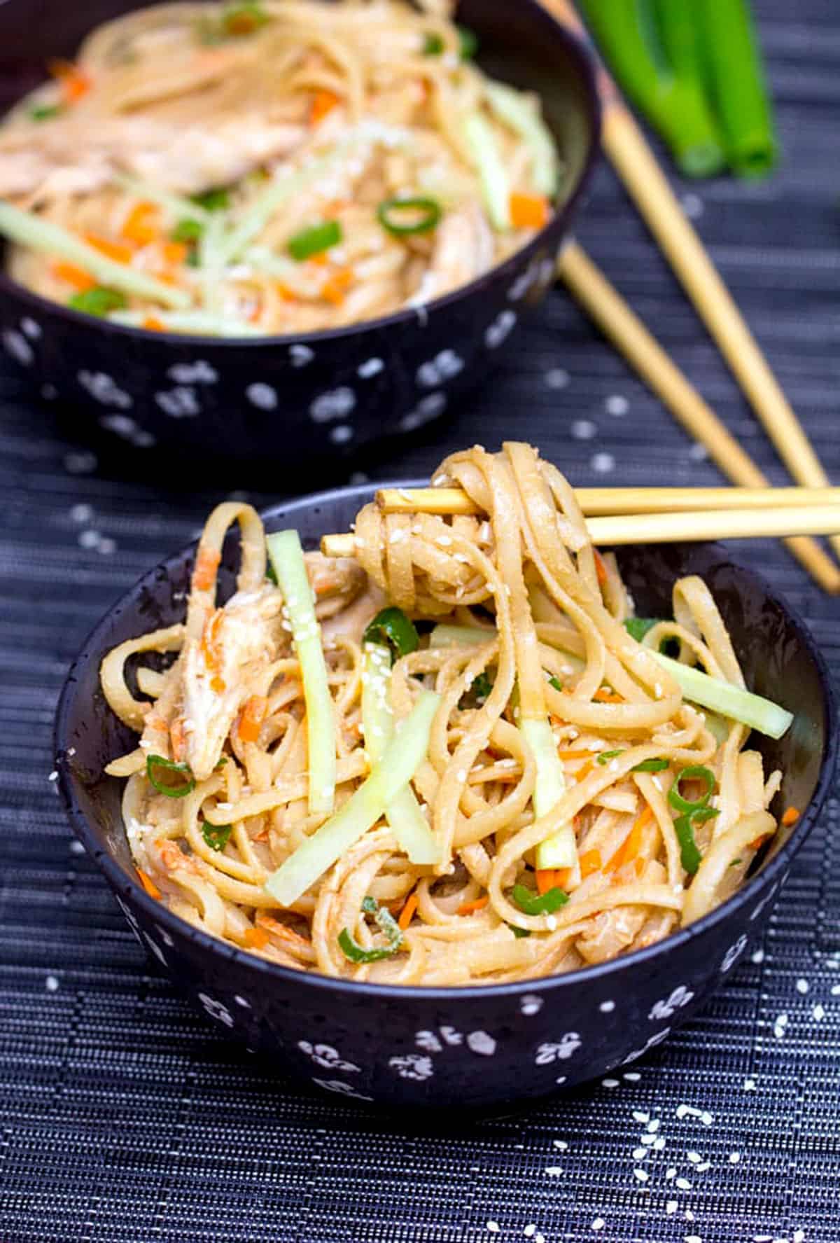 Black bowl filled with linguine coated with peanut sauce, bits of chopped carrots, scallions and sliced cucumbers. Chopsticks twirling some of the noodles.