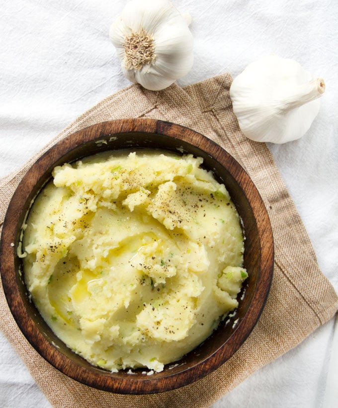  olive oil and roasted garlic mashed potatoes in a brown wooden bowl, on a tan dish towel, with two whole heads of garlic on the side