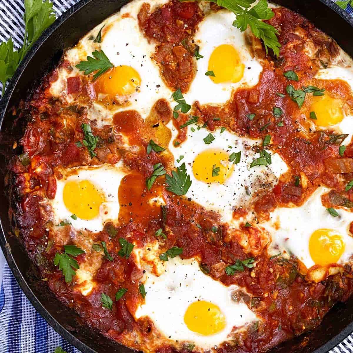 rich tomato sauce with peppers in a cast iron skillet topped with 7 cooked eggs and parsley leaves to garnish