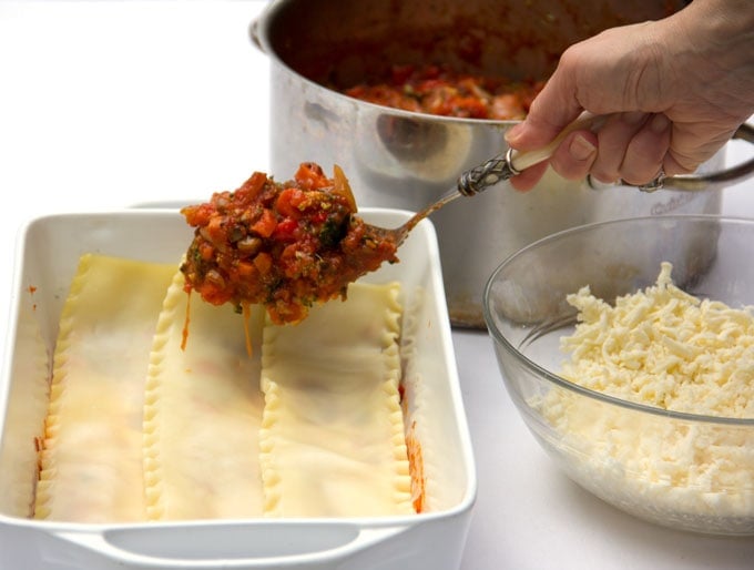 a white casserole dish with lasagna noodles lined up and a large spoon heaped with sauce about to be poured on top