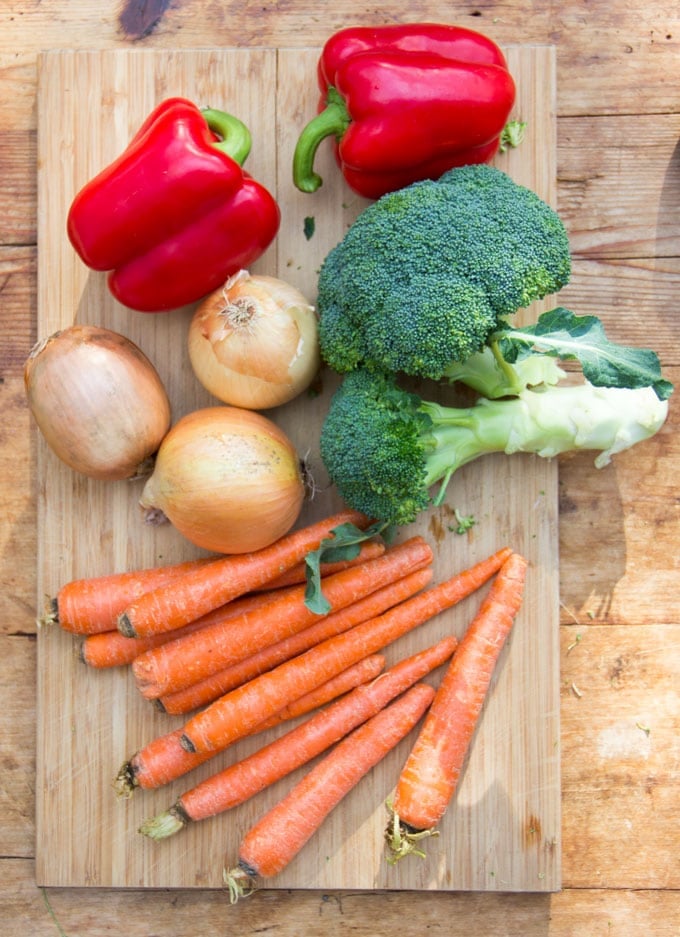 2 red bell peppers, 3 yellow onions, 1 bunch of broccoli and 10 carrots on a wooden cutting board.