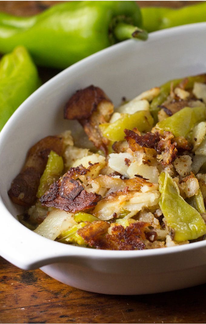 A white oval baking dish filled with crispy Italian fried potatoes and peppers, in the background area few green Cubanelle peppers