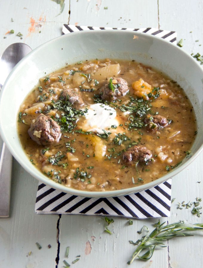 Light green bowl on a black and white striped napkin, filled with meatballs soup and garnish of sour cream and chopped herbs.