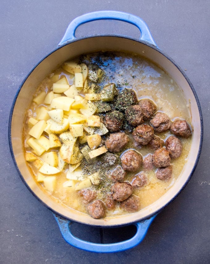 Blue Dutch oven filled with broth, cooked meatballs, diced potatoes, herbs and spices