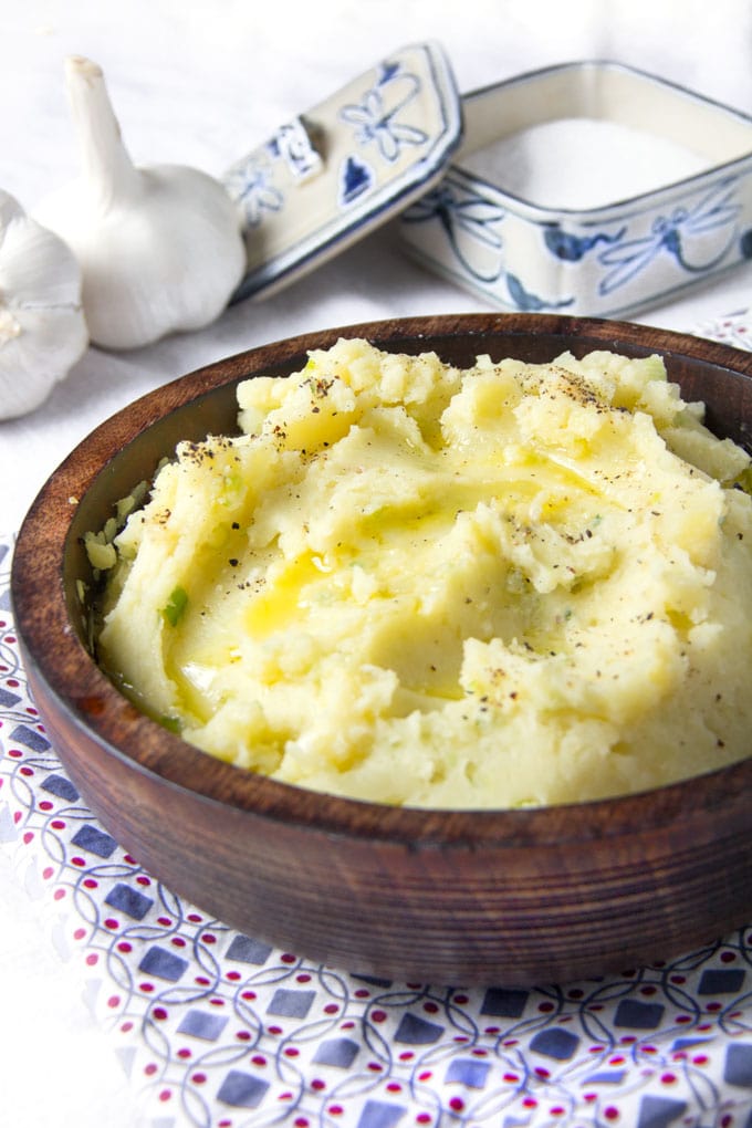 Brown wooden bowl filled with mashed potatoes. Two whole garlic bulbs in the background and a blue and white chine salt box in the background.