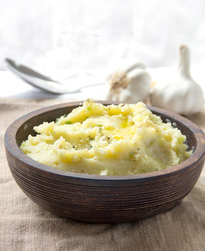 Brown wooden bowl filled with mashed potatoes. Two whole garlic bulbs in the background.