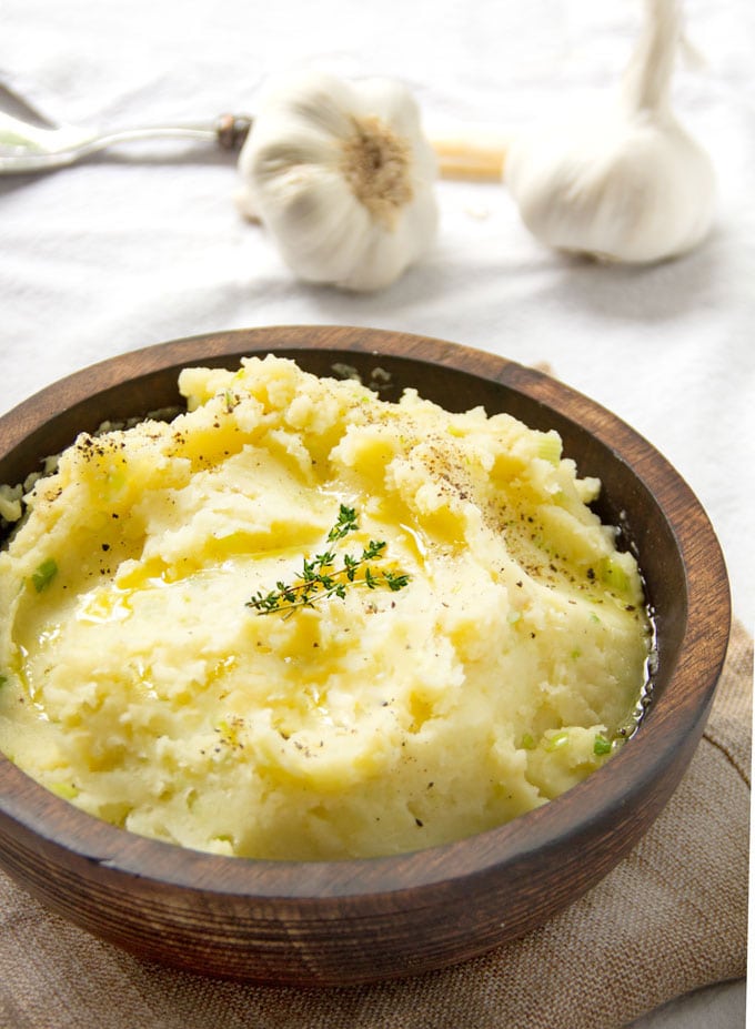 Brown wooden bowl filled with mashed potatoes and garnished with a sprig of thyme. Two whole garlic bulbs in the background.