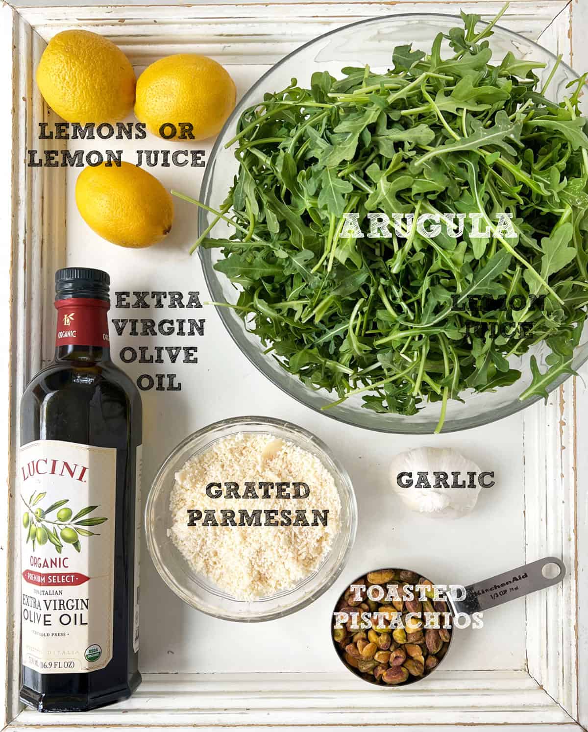 Ingredients for arugula pesto, seen from above: a bowl of arugula, three lemons, a bottle of olive oil, a small bowl of grated parmesan, some toasted pistachios and a bulb of garlic