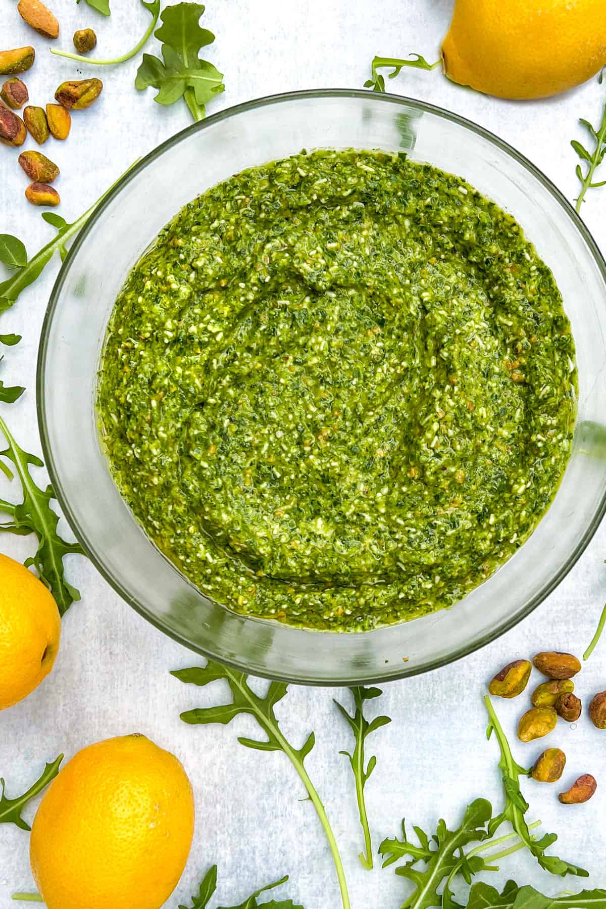 overhead shot of a glass bowl filled with arugula pesto, on a white countertop, surrounded by arugula leaves, 3 small piles of toasted pistachios and 3 lemons