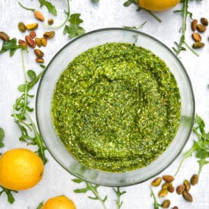 overhead shot of a glass bowl filled with arugula pesto, on a white countertop, surrounded by arugula leaves, 3 small piles of toasted pistachios and 3 lemons