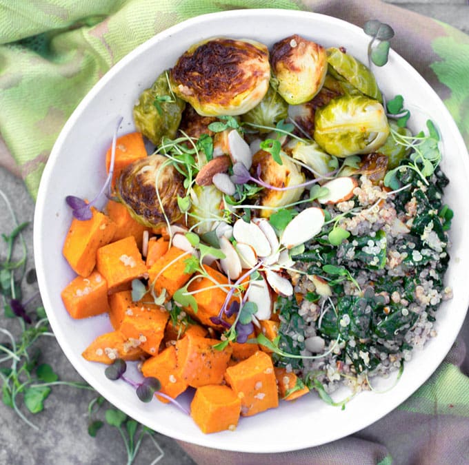 A buddha bowl on a green cloth napkin: A sweet potato Brussels sprouts buddha bowl with quinoa, wilted kale and a sprinkle of slivered almond and micro greens