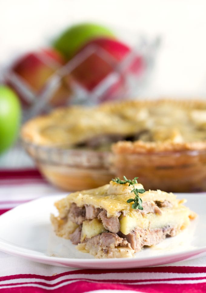 slice of cheshire pork pie in the foreground with the whole pie and a bowl of green and red apples in the background.