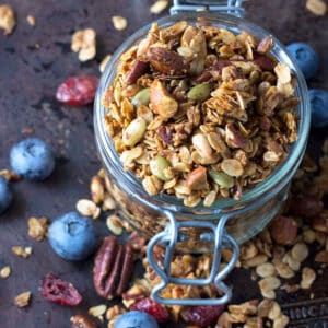 uncovered mason jar brimming and overflowing with nut and seed granola plus a few blueberries scattered around.