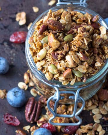 uncovered mason jar brimming and overflowing with nut and seed granola plus a few blueberries scattered around.