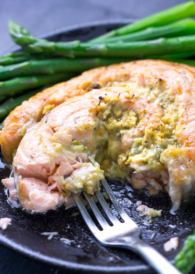 Black plate with a horseradish mustard stuffed salmon roulade and a fork stuck into it, with asparagus on the side.