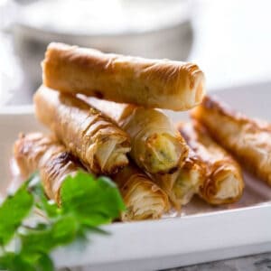 8 sigara borek rolls piled on on a small serving plate with a small bowl of dipping sauce in the background.