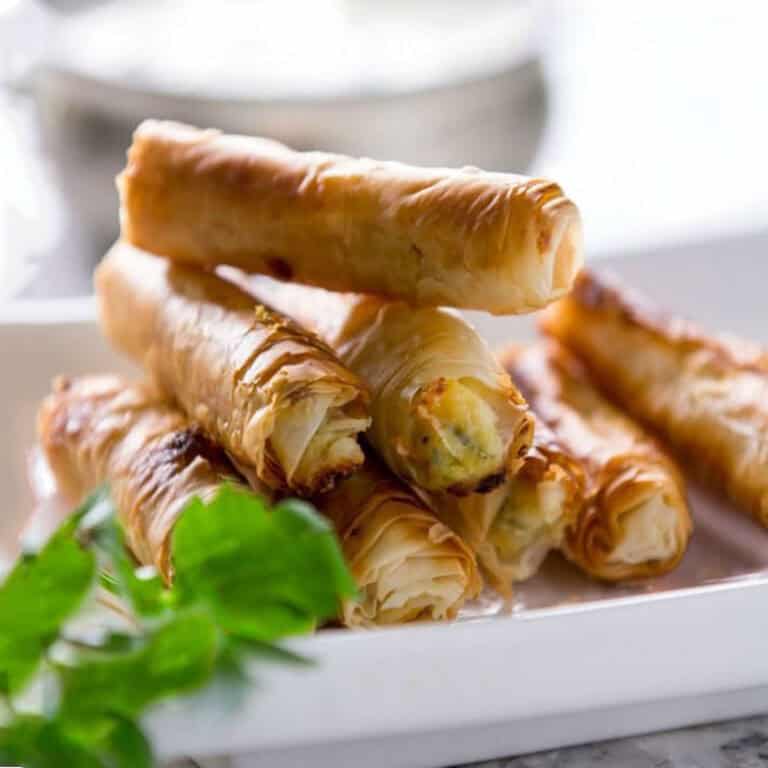 8 sigara borek rolls piled on on a small serving plate with a small bowl of dipping sauce in the background.