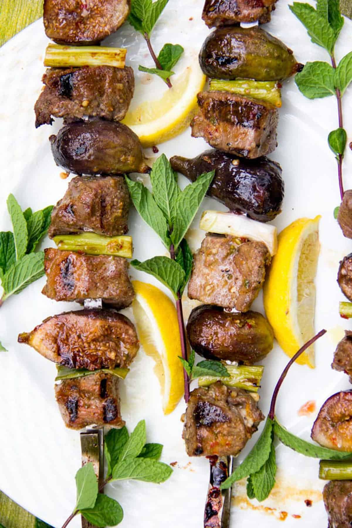 Two kebabs threaded with cubes of lamb, halved fresh figs and pieces of scallions, surrounded by lemon slices and mint sprigs
