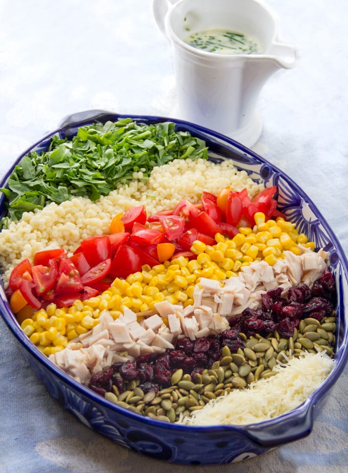 A chopped salad in an oval bowl, arranged on a rainbow of stripes, arugula, couscous, tomatoes, corn, turkey, cranberries, pumpkins seeds and cheese.