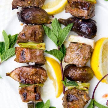 Two grilled lamb and fig skewers surrounded by lemon slices and mint sprigs